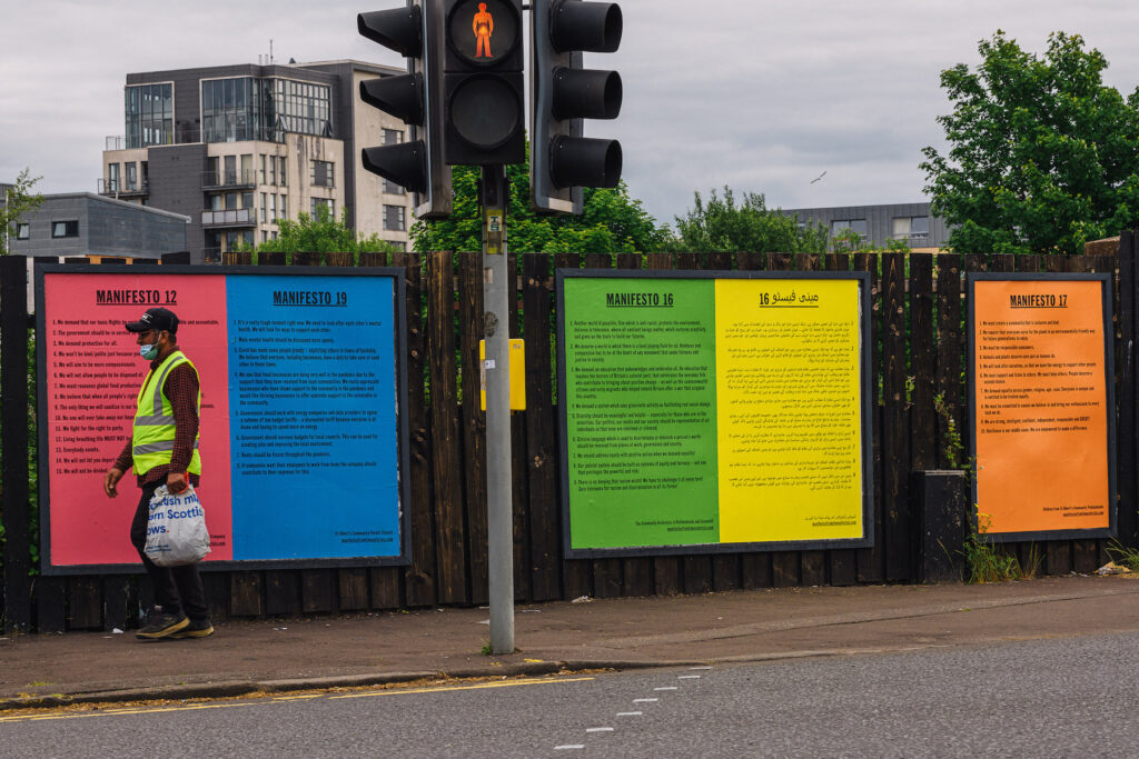Man in hi-vis vest, wearing face mask walking past billboard posters of five manifestos in Pollokshield, Glasgow
